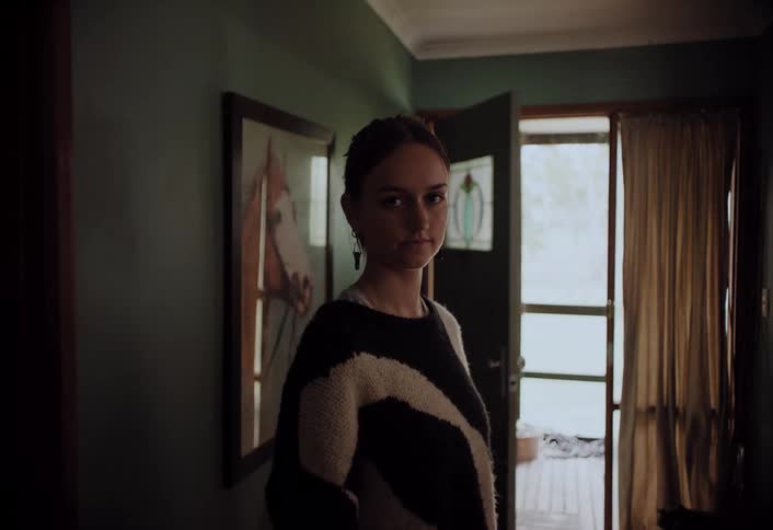 A woman with dark hair pulled back stands in a dimly lit hallway looking directly at the camera with a neutral expression