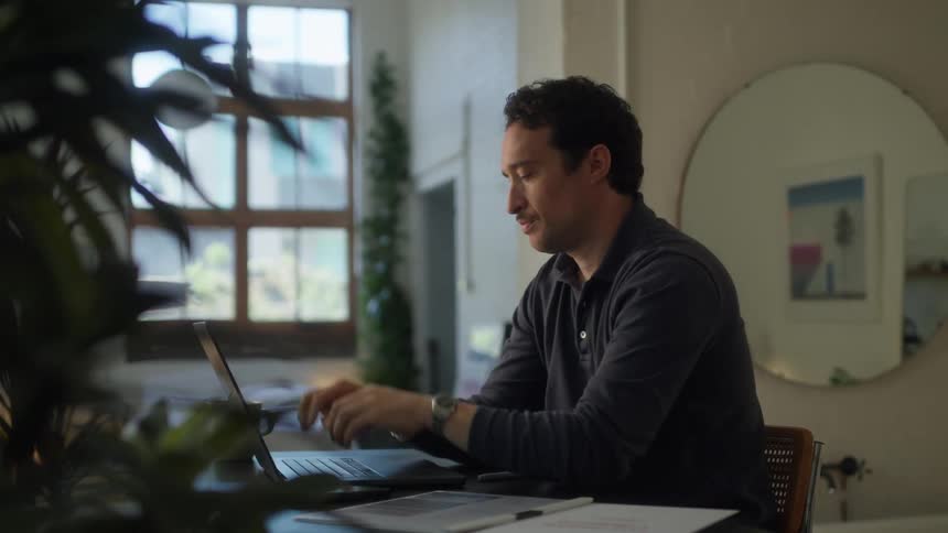 A man with dark curly hair sits at a desk in a well lit room looking thoughtfully into the distance with his hands clasped under his chin before turning his attention to type on a laptop