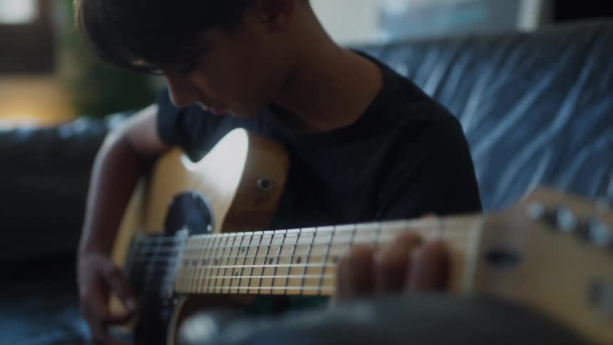 A boy sits on a couch practicing electric guitar while an adult assists him at home