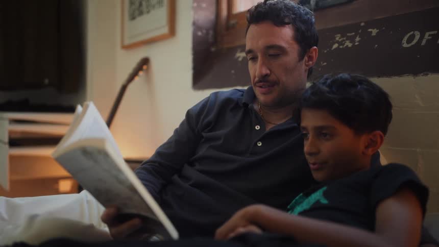 A father and son sit together in bed reading a book in a cozy bedroom