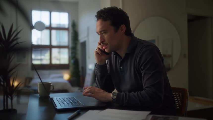 A man is using a laptop while talking on a cell phone