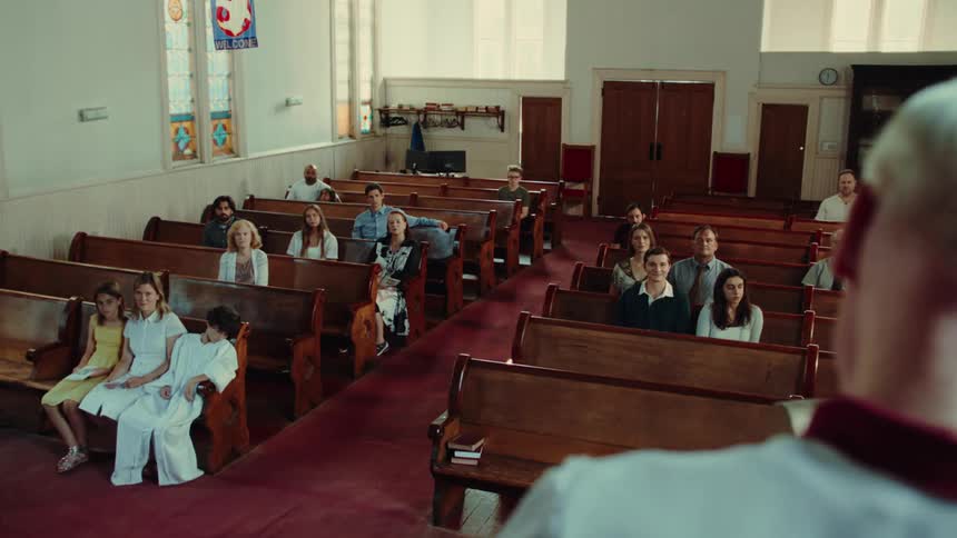 A small congregation of people including families and individuals sits on wooden pews inside a modest church attentively facing a man leading a service at the front