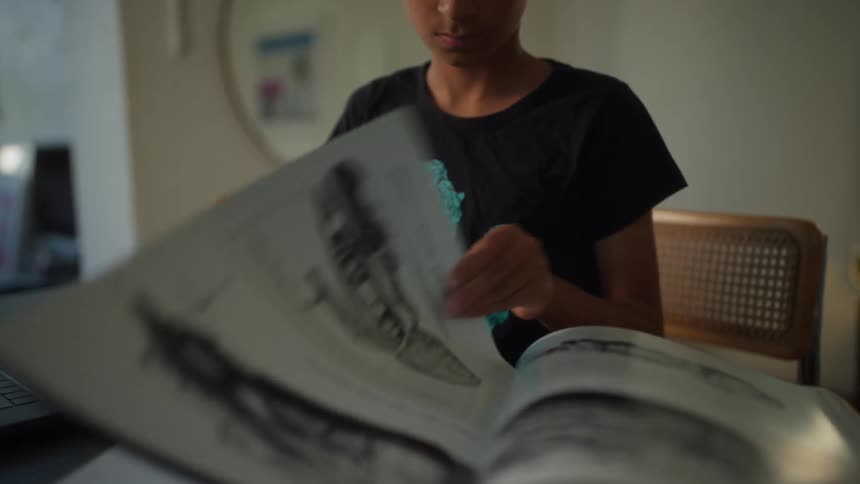 Youth examines a book before turning to his computer while sitting at home during the daytime