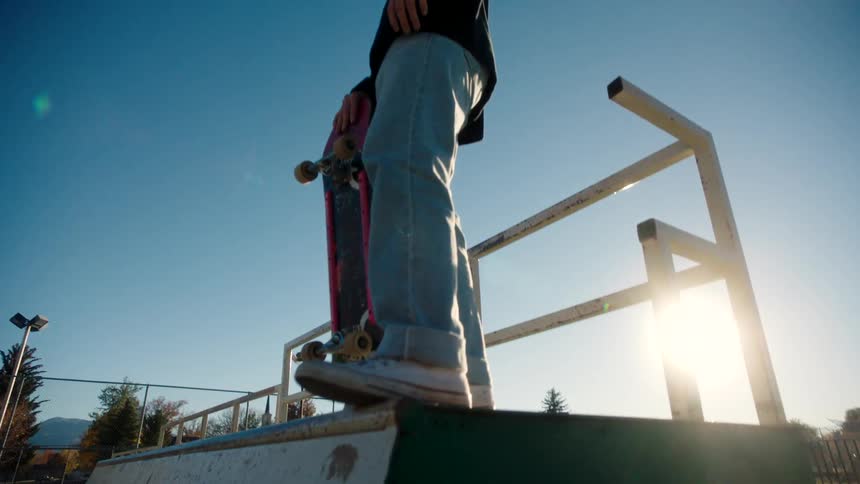 A teenage male wearing loose blue jeans and a long sleeve black shirt holds a skateboard as he stands on top of a wooden skate ramp flanked by metal piping under a clear blue sky