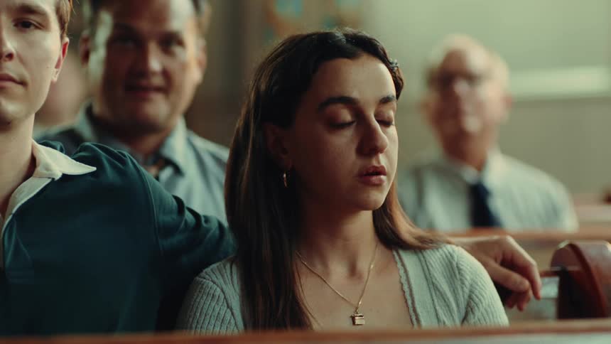 A young woman with long brown hair wearing a light gray cardigan over a white top and a delicate necklace is looking slightly to her right with a concerned expression while sitting in a row with other people in a church