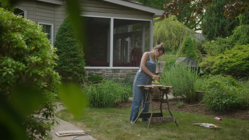 A woman in overalls sands a wooden table in the front yard of a home at morning