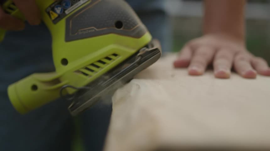 A man is using a light green belt sander to smooth out the edge of a piece of wood while he uses his left hand to stabilize it