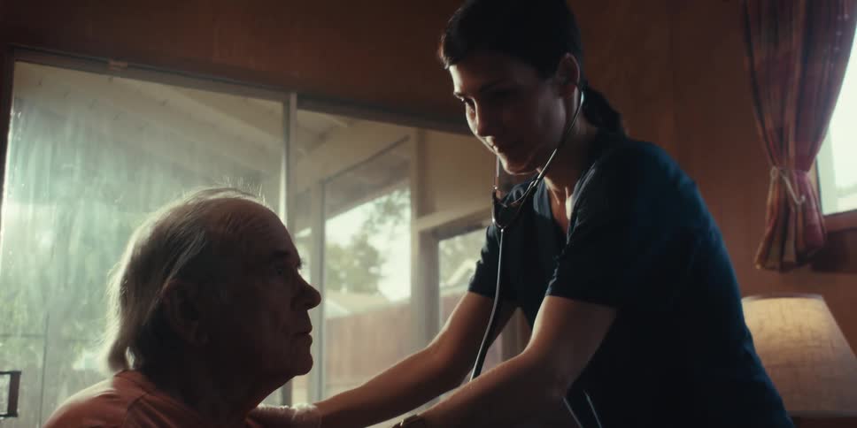 A nurse with a stethoscope tends to an elderly man sitting on a couch at home