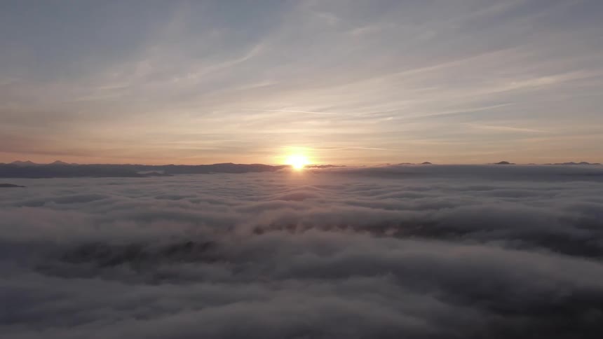 Aerial view of a thick layer of clouds stretching to the horizon with the sun just breaking through casting a warm golden glow on the cloud tops under a soft partly cloudy sky