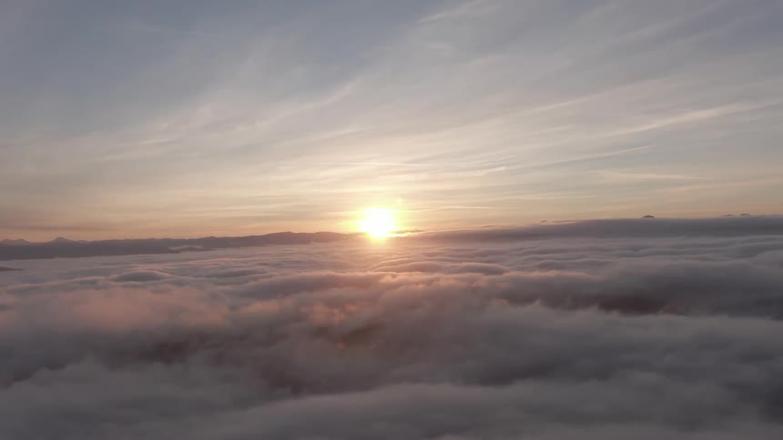 The sun rises above a sea of clouds casting warm light across the sky as an aerial view floats gently through a soft glowing morning atmosphere