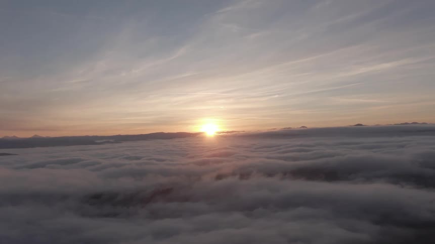 Aerial view of a thick layer of clouds stretching to the horizon with the sun just breaking through casting a warm golden glow on the cloud tops under a soft partly cloudy sky