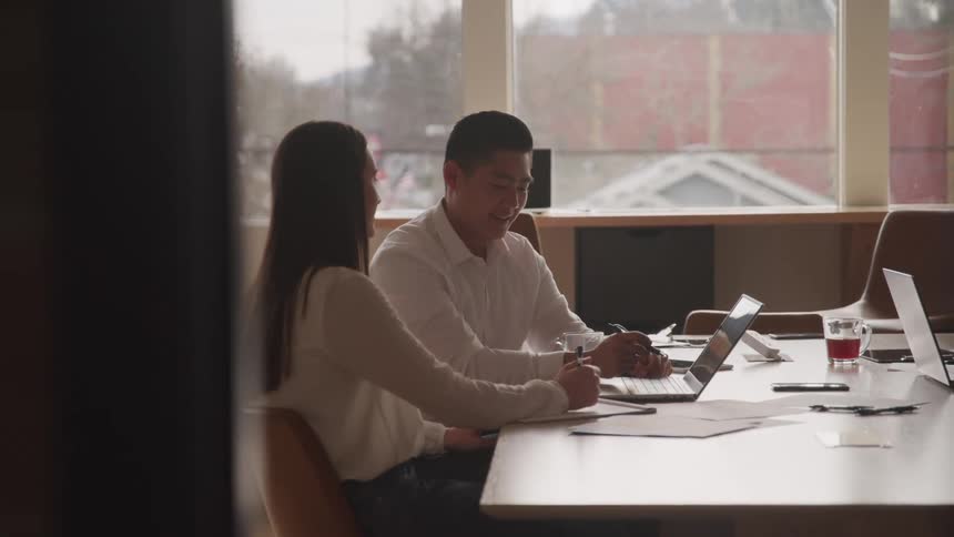 Two people work at an office desk talking while looking at the same laptop in a welllit room with large windows