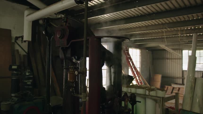 An industrial workshop features a large steaming engine with sunlight streaming through the windows