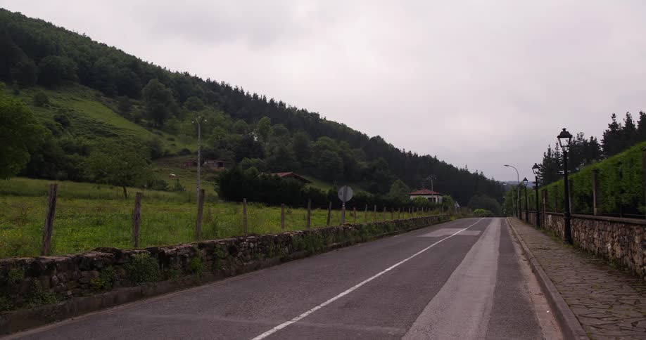 A quiet twolane road runs through a rural valley as large plants behind stone walls sway gently in the breeze and clouds above drift slowly across the overcast sky
