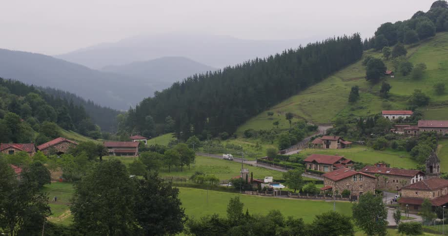 A picturesque rural landscape with houses and buildings scattered across lush green fields and rolling hills framed by dense forests and mountains