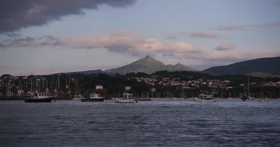 Several boats are moored in a tranquil harbor surrounded by a coastal town and green hills with a towering mountain under a partly cloudy sky