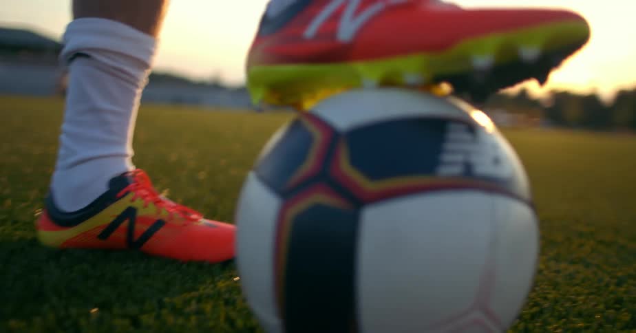 Man wearing bright orange cleats rolling around a soccer ball at sunset