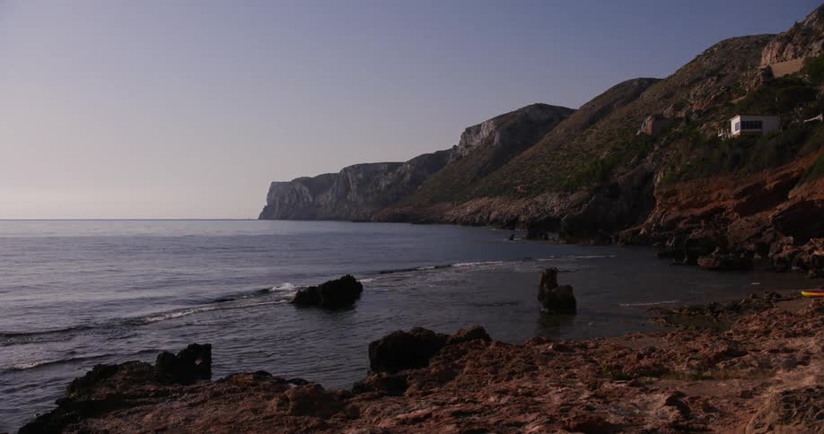 A rocky coastline meeting a calm dark blue sea under a clear pale sky with imposing cliffs rising along the shore