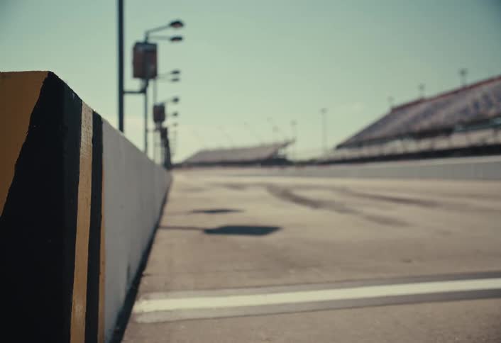A blurred view down a paved racetrack shows tire marks and grandstands under a hazy sky with a black and yellow striped barrier in the foreground