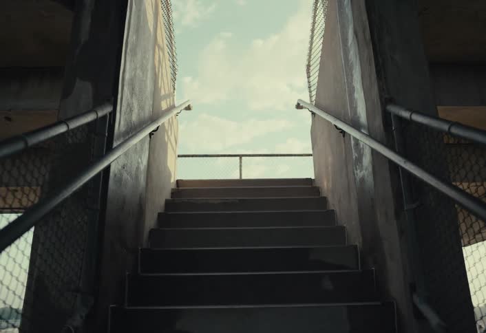 A concrete staircase with metal railings leading upwards towards an opening that reveals a view of a flat landscape under a cloudy sky