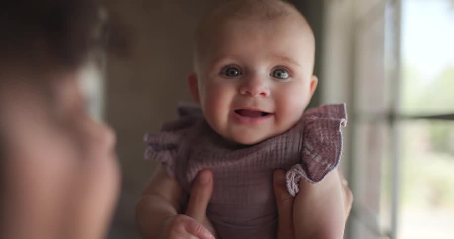 A baby with fair skin and light eyes wearing a purple outfit is held up by an adult looking towards the camera with a happy expression