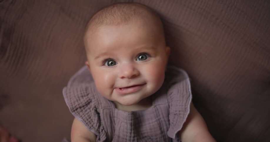 A baby with fair skin and light eyes wearing a textured purple outfit lying on a brown surface