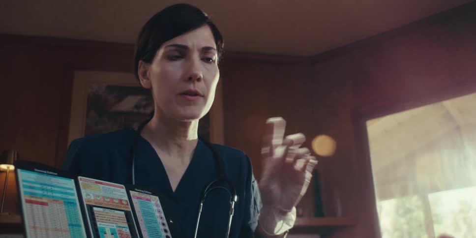 A nurse wearing gloves with a stethoscope around her neck talks to a patient at home while holding a medical chart