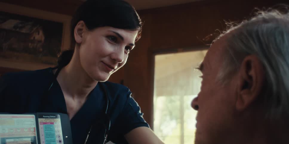 A healthcare professional shows a medical chart to an elderly man in a cozy room with a fireplace in the background