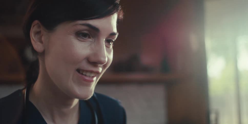 A closeup of a nurse with a stethoscope over her shoulders smiling warmly as she tends to someone at home