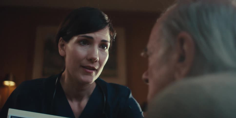 A healthcare professional with dark hair wearing a dark scrub top and holding a medical image is looking and speaking to an older patience