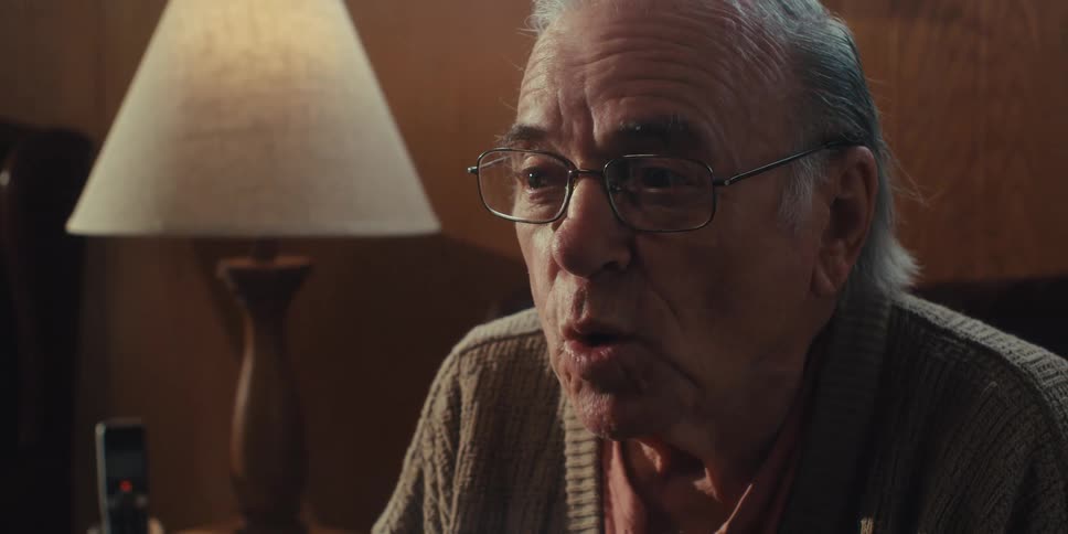 An elderly man smiles as he talks and sits on his couch