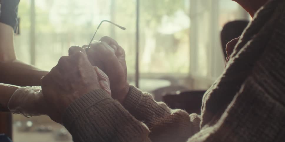 A nurse with a stethoscope hanging around her neck holds both hands of an elderly man sitting on a couch at home