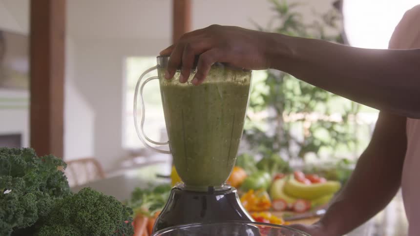 A person prepares fresh green vegetable juice using a blender in a brightly lit home kitchen