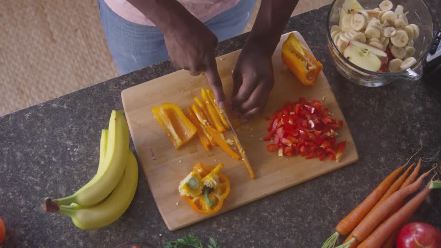 A man is slicing yellow bell peppers on a wooden cutting board surrounded by chopped red peppers whole carrots tomatoes bananas and a bowl of sliced bananas on a dark countertop