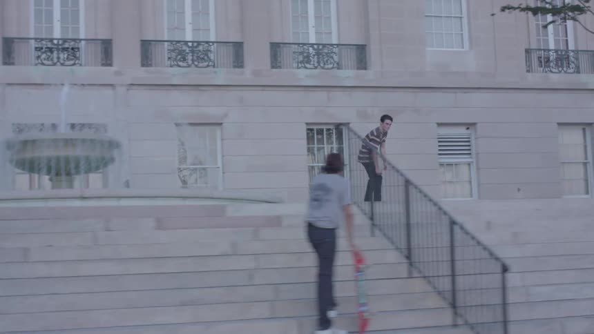 A skateboarder performing a trick off a staircase in front of a large building observed by two other people