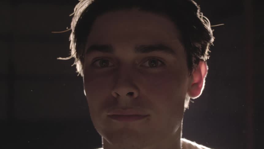 A closeup of a young male looking directly at the camera with bright lamp light illuminating his head from behind