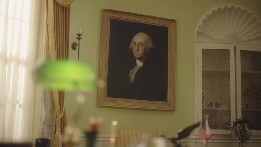 A framed portrait of a person in a white wig and dark clothing hangs on a wall in a classically decorated room with a green desk lamp a candle holder and a glassfronted cabinet