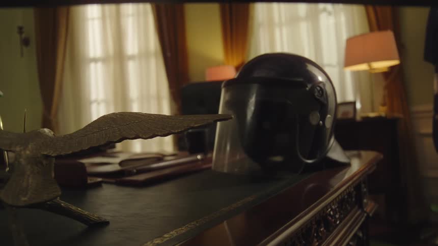 A wooden desk is adorned with an american flag an eagle figurine and a police riot helmet situated in a warmly lit room with large windows and heavy curtains