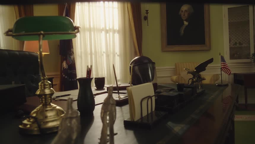 A traditional office features a large desk adorned with a green lamp an eagle statue and an american flag with a portrait hanging on the wall and curtained windows in the background