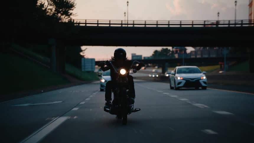 A motorcyclist rides along a highway at dusk flanked by cars with an overpass and city skyline in the background