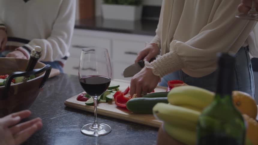 Hands of a woman with a knife cutting vegetables on a wooden board on a countertop with a glass of wine