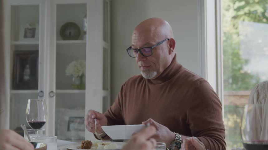A person serves food from a bowl onto their plate at a dining table surrounded by others