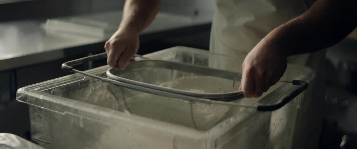 A man uses a mesh strainer to sift flour into a large clear plastic container in a commercial kitchen