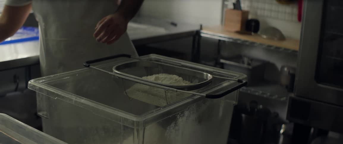 A man pours flour from a glass measuring bowl and then sifts it through a mesh strainer into a large plastic container inside a stainless steel commercial kitchen during food preparation