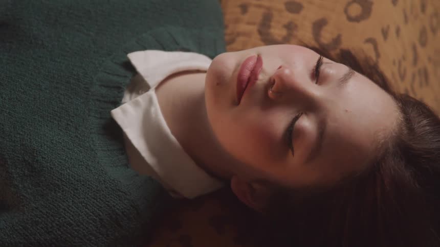 A close up shows a young girl with delicate features wearing a green sweater over a white collared shirt resting on a bed