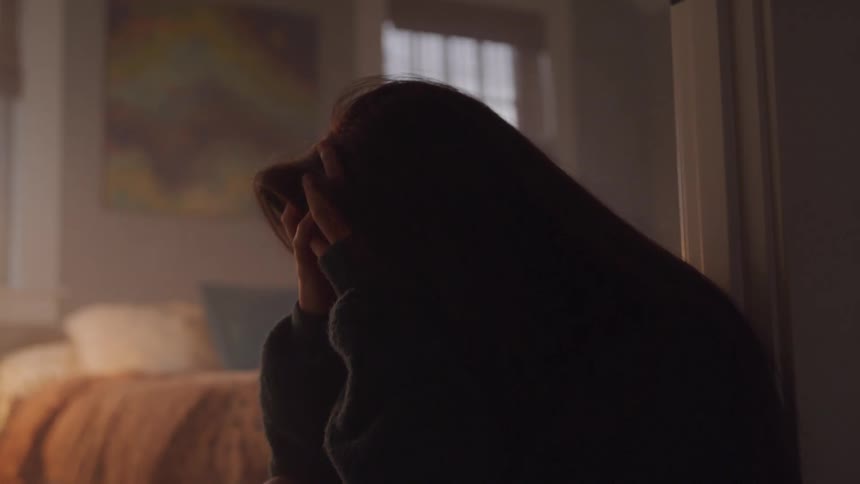 A teenage girl sits in a dimly lit room holding her head in both hands conveying sadness or distress