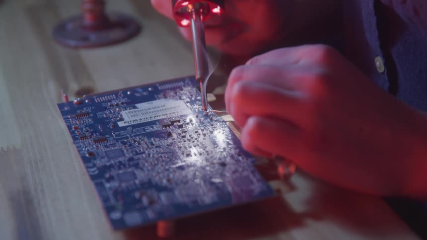A person solders components onto a computer graphics card at a wooden desk using a precision tool under red led lighting during a handson electronics repair session