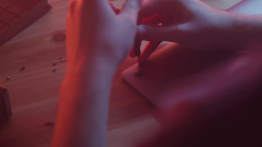 Hands use a small screwdriver to carefully remove tiny screws from the back cover of a macbook laptop on a wooden surface