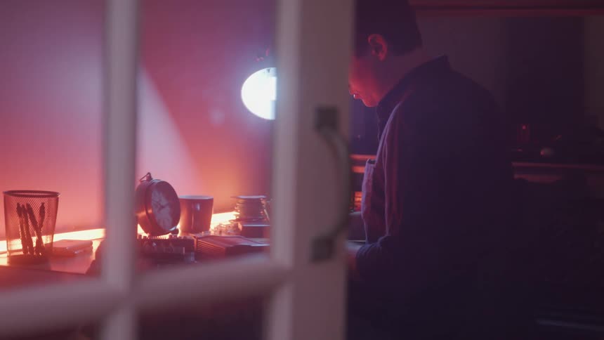A person carefully assembles an electronic device at a desk illuminated by a bright lamp with various tools and components spread around