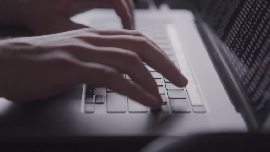 A close up of hands typing on a silver laptop with visible coding displayed on its screen
